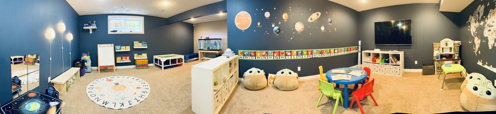 The dayhome playroom end to end, with a reading corner, art easel, sensory table, solar system wall, and a large world map